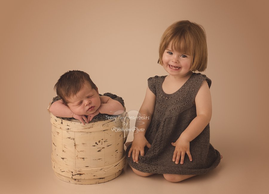 søskende foto med storesøster i grå kjole og nyfødt bror i spand ved siden af.